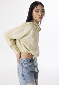  Lichtgele blouse met kraag en een gestructureerde afwerking, gecombineerd met versleten blauwe jeans met bedrukte accenten.