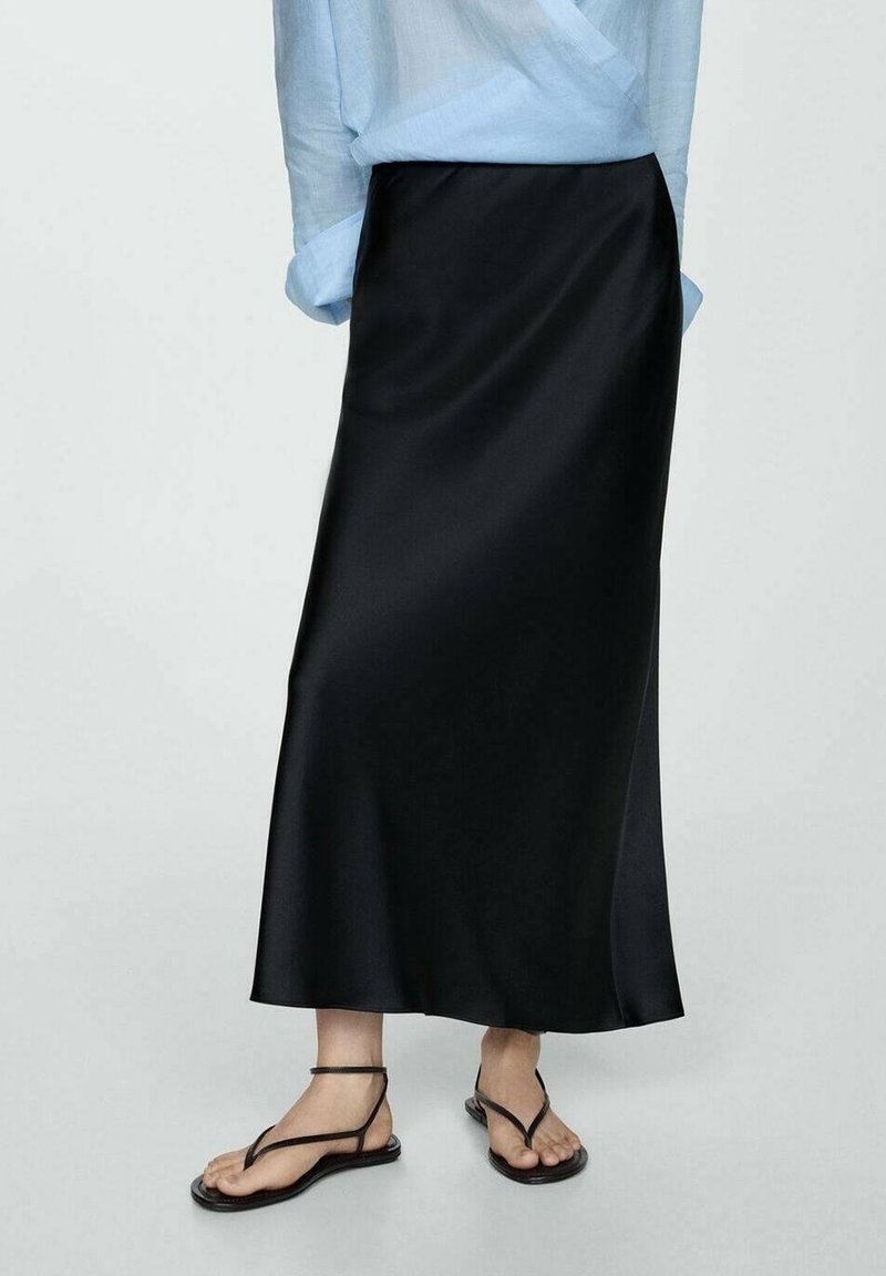 Femme portant une jupe midi en satin noire associée à un chemisier bleu clair et des sandales plates noires à brides sur un fond uni.