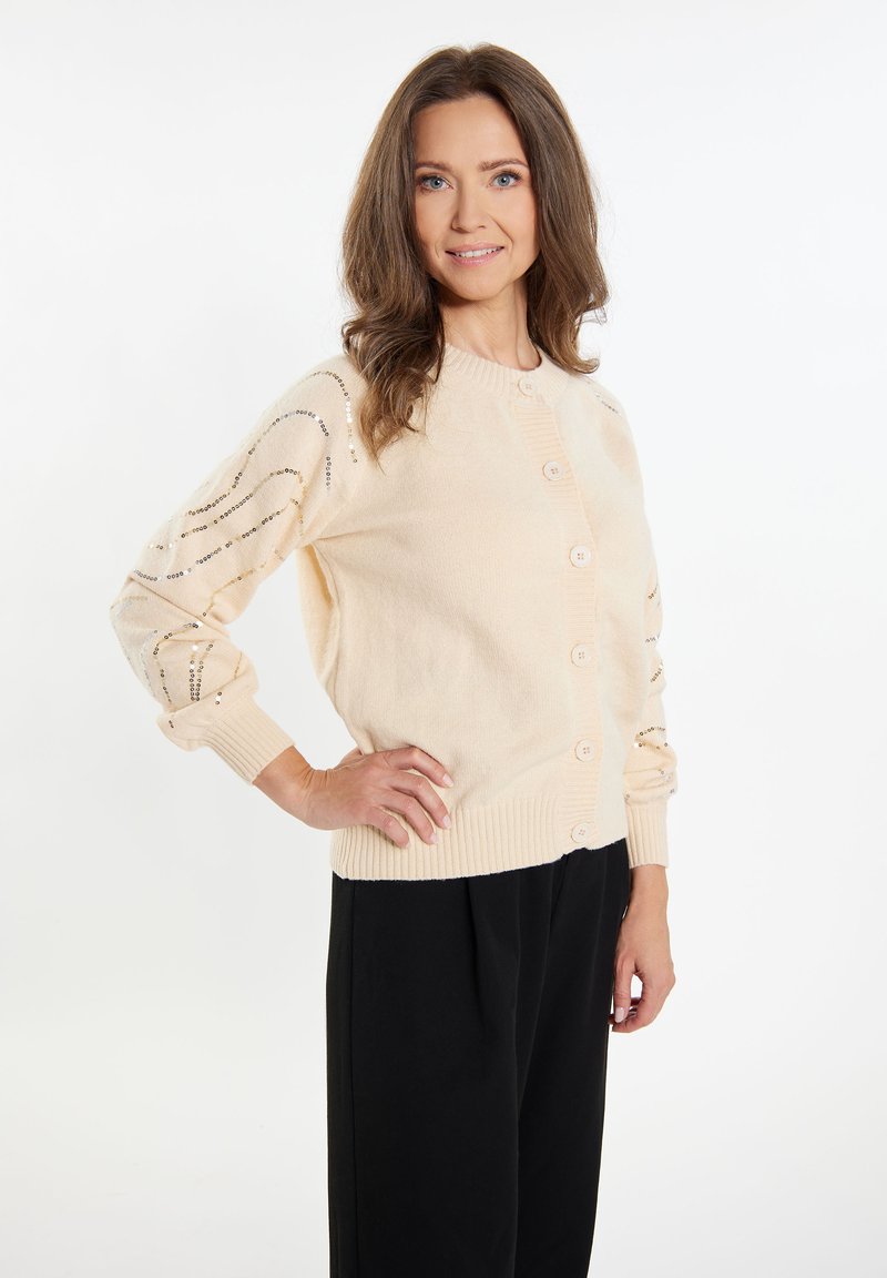 Cardigan beige chiaro realizzato in materiale morbido, con colletto rotondo, chiusura a bottoni e dettagli decorativi con paillettes sulle maniche.