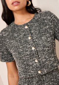 Mustvalge tweedist lühikeste varrukatega top, millel on kuldsed nööbid ees, tekstureeritud kangas ja ümmargune kaelus.