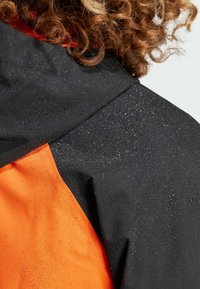 Sort og orange vandtæt jakke med vanddråber på stoffet. Har tydelig farveblokeringsdesign og en struktureret overflade.