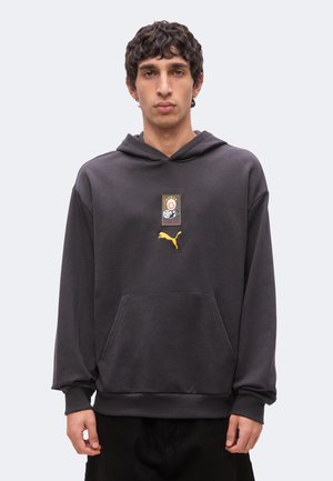 Jeune homme portant un sweat à capuche gris foncé avec une poche avant et un logo Puma sous un petit motif sur la poitrine, debout devant un fond uni.