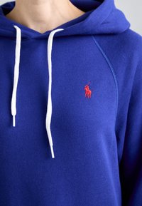 Kék kapucnis felső puha anyagból, kenguruzsebes kialakítással, fehér zsinórokkal és egy kis piros hímzett logóval a mellkason.