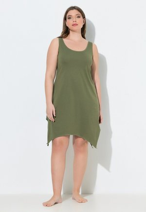 Woman stands barefoot against white wall, wearing sleeveless olive green asymmetrical hem dress.