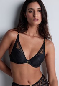 Soutien-gorge en dentelle noire avec des bonnets triangulaires, arborant un motif floral, des accents en métal doré et des bretelles réglables. Texture lisse.
