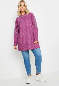 Lila tröja med oversized passform och ribbat halslinning, med metalliska detaljer på ärmarna; kombinerad med ljusblå skinny jeans och vita sneakers.