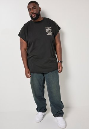 Uomo in piedi davanti a uno sfondo semplice che indossa una maglietta nera senza maniche, jeans blu larghi, sneakers bianche e un orologio da polso.