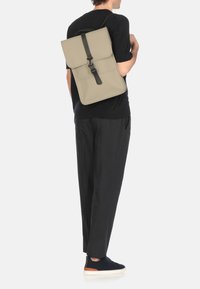 Zaino in materiale beige chiaro con chiusura a patta e tracolla nera. Progettato con caratteristiche minimaliste, forma rettangolare di media grandezza.