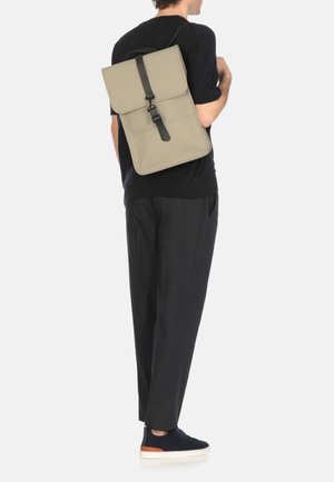 Mochila de material beige claro con cierre de solapa y correa negra. Diseñada con características minimalistas, de forma rectangular mediana.
