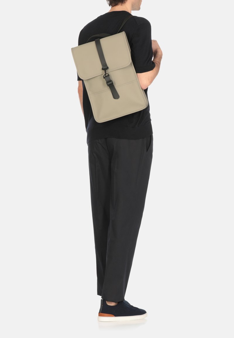 Zaino in materiale beige chiaro con chiusura a patta e tracolla nera. Progettato con caratteristiche minimaliste, forma rettangolare di media grandezza.