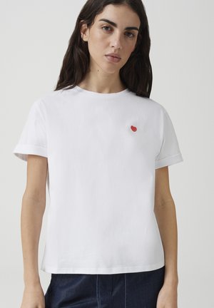Femme aux longs cheveux foncés portant un t-shirt blanc uni avec un petit détail en forme de cœur rouge sur le côté gauche de la poitrine, debout devant un fond neutre.