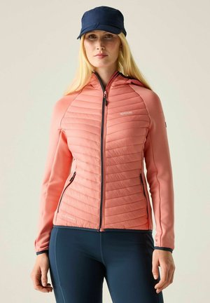 Mujer con chaqueta acolchada coral con cremallera y bolsillos, leggings azul marino y gorra azul marino, mirando hacia adelante sobre un fondo liso.