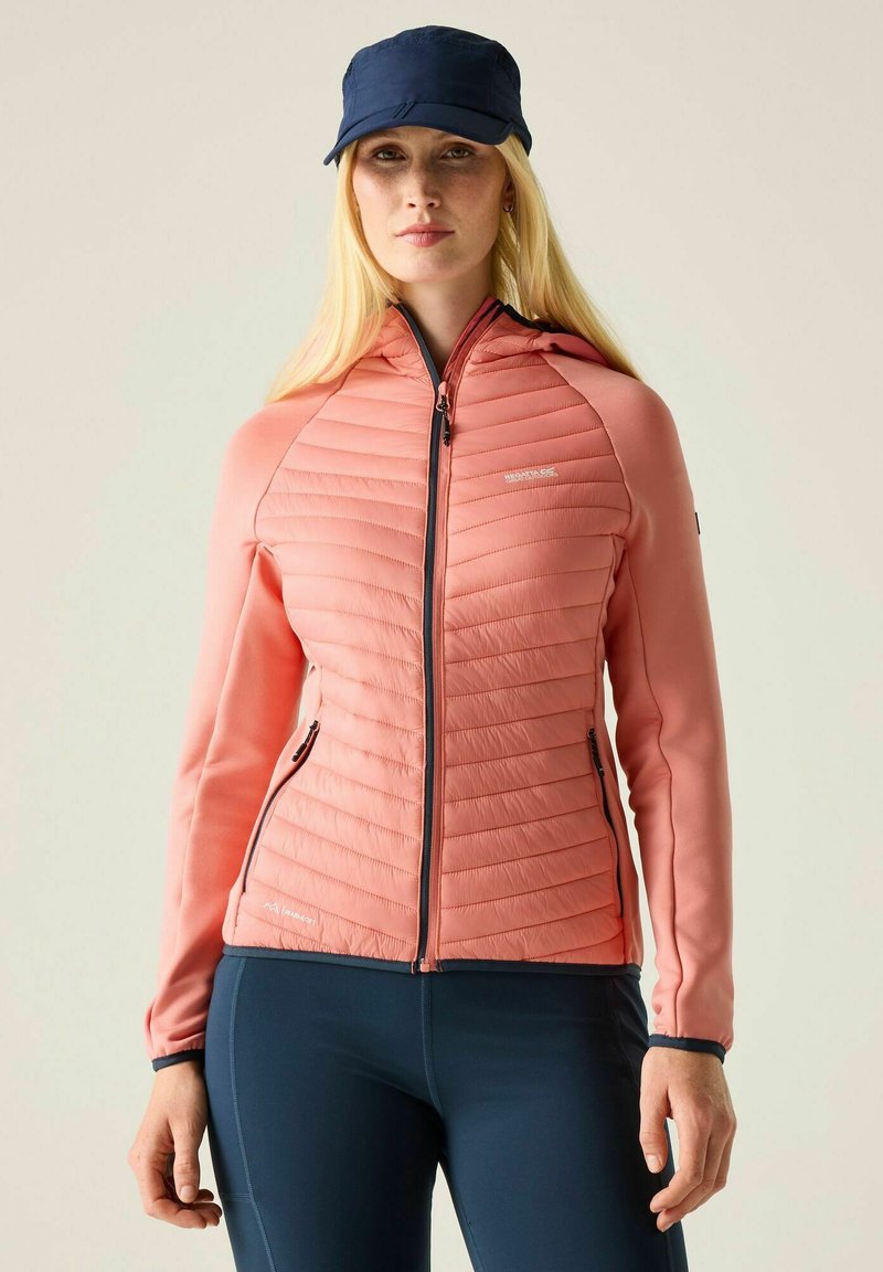 Femme portant une veste matelassée corail zippée avec poches, leggings bleu marine et casquette bleu marine, tournée vers l'avant sur un fond uni.