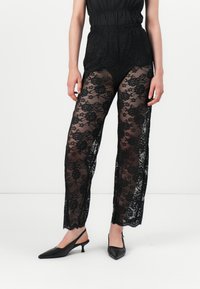 Pantalons en dentelle noire avec un motif floral et des sections transparentes. Dotés d'une taille haute et d'une coupe décontractée, associés à des escarpins noirs à talons ouverts.