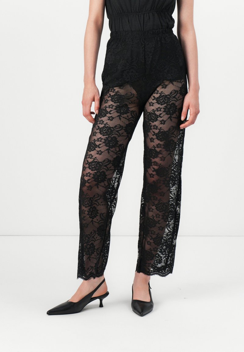 Pantalons en dentelle noire avec un motif floral et des sections transparentes. Dotés d'une taille haute et d'une coupe décontractée, associés à des escarpins noirs à talons ouverts.