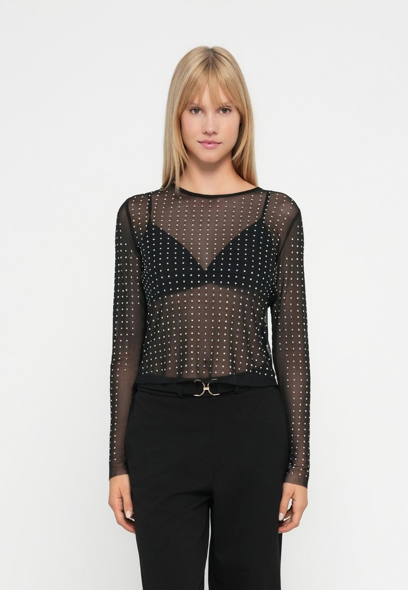 Fekete hálós, hosszú ujjú top fémpántokkal díszítve, áttetsző dizájnnal és testhezálló siluettel. Fekete bralette felett viselve.