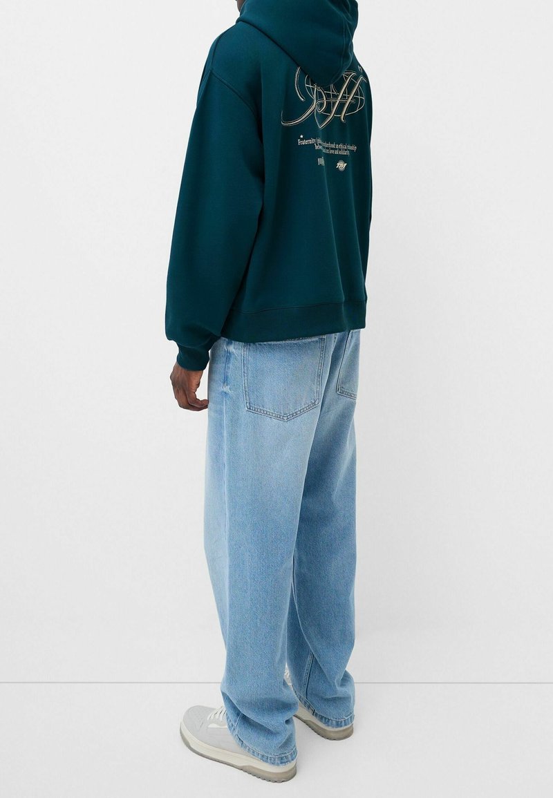 Personne portant un sweat à capuche vert foncé avec un texte dans le dos, un jean large bleu clair et des baskets blanches, debout contre un fond clair uni.