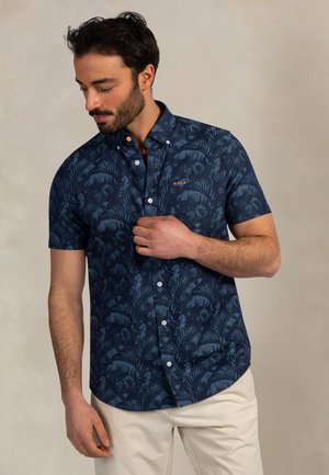 Man met donker haar en baard die een knoop aanpast op een marineblauw, kort mouwen overhemd met patroon, draagt lichtbeige broek, staand tegen een effen achtergrond.