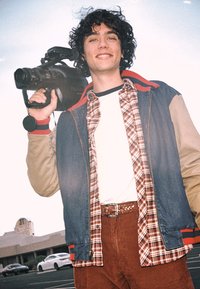 Person holding a video camera, wearing a denim jacket with red and beige accents over a plaid shirt and white t-shirt, paired with brown corduroy pants.