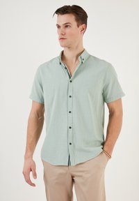 Cămașă cu mâneci scurte, de culoare verde deschis, dintr-un material texturat, cu guler cu nasturi, având nasturi închis la culoare și un tiv curbat.