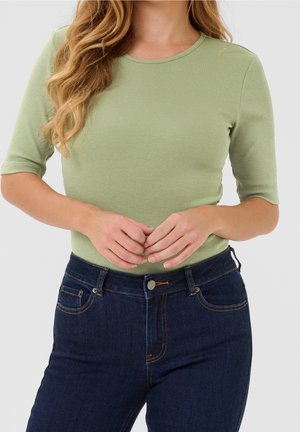 Vrouw met golvend haar die een lichtgroene aangesloten geribde top met ellebooglange mouwen en donkerblauwe hooggetailleerde spijkerbroek draagt, handen voor zich geklemd.