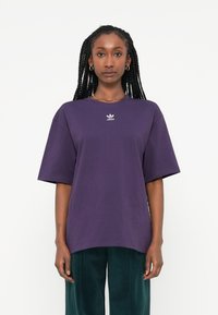Violetti puuvillainen t-paita lyhyillä hihoilla, jossa on pieni valkoinen Adidas-logo rinnassa ja rento leikkaus. Rakoileva kangas.