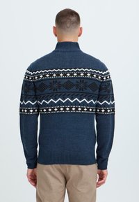 Tamsiai mėlynas megztinis su aukštu apykakle, turintis raštuotas juostas juodos, baltos ir mėlynos spalvų. Raukšlėti rankogaliai ir apatinė dalis, tekstūrinis paviršius.