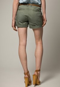 Pantaloni shorts verde oliva con risvolti, due tasche posteriori e una cintura marrone con dettagli perforati. Indossati con sandali col tacco alti color carne.