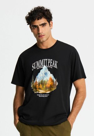 Uomo con capelli ricci scuri che indossa una maglietta nera con una grafica di montagna e alberi e testo "Summit Peak Nature Experience."
