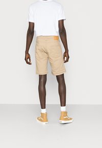 Hombre vistiendo pantalones cortos beige, camiseta blanca, calcetines blancos y zapatillas altas marrón, de pie de espaldas sobre un fondo gris liso.