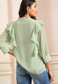 Bluză semi-transparentă verde deschis, cu accente de volănașe și buline texturate, având mâneci lungi și un tiv curbat. Poartă cu blugi albaștri.
