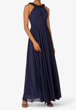 Kobieta w granatowej, bezrękawnikowej sukni sięgającej podłogi z kwiatowym zdobieniem przy dekolcie i plisowaną talią, ubrana w czarne, odkryte na palcach szpilki.