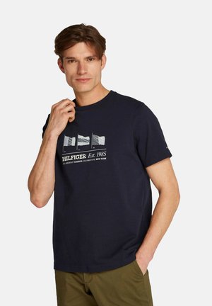 Camiseta de algodón color azul marino con mangas cortas. El frontal presenta un gráfico de banderas y el texto "HILFIGER Est. 1985" en blanco. Textura suave.