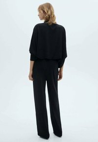 Black, textured wide-leg jumpsuit with a loose-fitting, cropped top and long sleeves. Simple design and monochromatic color scheme.
