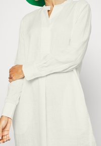 Tunique en lin blanc à manches longues, dotée d'un col cranté et de poignets boutonnés, texture lisse et coupe ample.