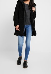 Abrigo de forro polar negro con capucha, bolsillos frontales, cierre mediante cremallera; camiseta gris debajo, jeans skinny azules y zapatos negros con cordones.
