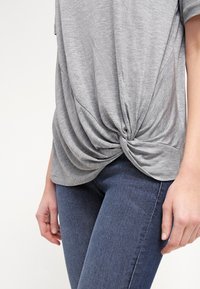 Gray top featuring a knotted front detail, short sleeves, and a soft, lightweight fabric, paired with dark blue fitted jeans.