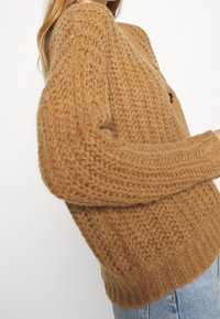 Brauner gestrickter Pullover mit strukturiertem Muster, runder Ausschnitt und seitlichem Knopfdetail. Locker sitzend, mit langen Ärmeln.