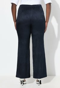 Marineblaue weite Textilhosen mit einem subtilen Wellenmuster, elastischem Bündchen und glänzenden silbernen High Heels auf einem weißen Hintergrund.