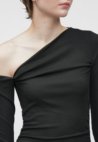 Μαύρη ριχτή μπλούζα μακρυμάνικη με διαγώνιο λαιμόκοψη, που διαθέτει φάσες και σχέδιο χωρίς ώμους. Λείο υλικό με εφαρμοστό σχήμα.