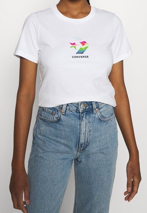 Osoba nosząca białą koszulkę Converse z kolorowym logo w połączeniu z jasnoniebieskimi dżinsami z wysokim stanem na jednolitym tle.