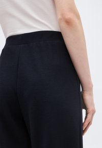 Άτομο που φοράει μαύρο παντελόνι ψηλόμεσο και λευκό κοντομάνικο πουκάμισο μπα tucked in, με το δεξί χέρι χαλαρό στο πλάι.