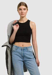Jeune femme aux cheveux blonds portant un crop top noir et un jean bleu clair, tenant une veste grise sur l'épaule, face à l'avant.