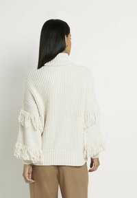 Pull crème en grosse maille avec un col haut, présentant des détails en franges texturées le long des manches et un ourlet côtelé.