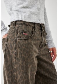 Osoba w brązowych spodniach w panterkę i jasnoszarym swetrze zapinanym na guziki, z dłonią spoczywającą w przedniej kieszeni spodni, ozdobiona srebrnymi pierścionkami.