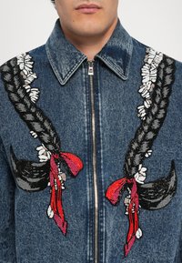 Giacca in denim con ricami neri intrecciati, dettagli floreali e nastri rossi sulla parte anteriore, colletto con bottoni e chiusura con zip.