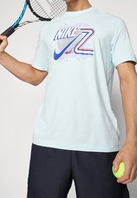 Světle modré tričko s grafickým potiskem s názvem "Nike" a logem tenisového míčku. Krátké rukávy, kulatý výstřih, spárováno s tmavými šortkami.