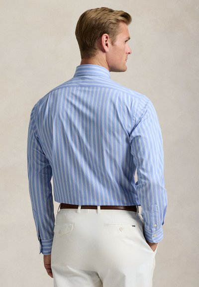 Ein Mann, von hinten gesehen, trägt ein blau gestreiftes Langarmhemd, das in weiße Hosen mit braunem Gürtel gesteckt ist, Hand in der Tasche.