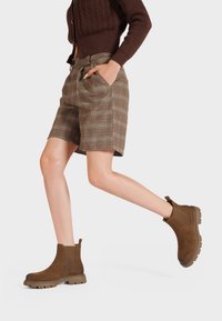 Stivaletti in suede marrone con pannelli elastici laterali e suola testurizzata, abbinati a pantaloni corti a quadri a vita alta. Modello in movimento.