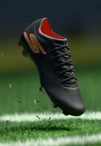 Cizma de fotbal neagră cu accente portocalii; partea superioară texturată, dantelă vizibilă și cu ținte modelate pe o suprafață de iarbă, suspendată în aer.